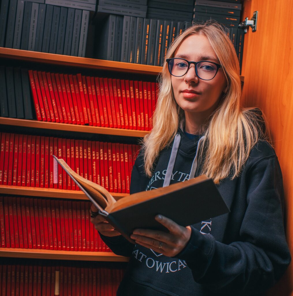 Młoda kobieta o długich blond włosach i w okularach stoi przed regałami wypełnionymi czerwonymi i czarnymi książkami, trzymając w ręku otwartą książkę i ubrana w ciemną bluzę z kapturem.