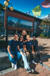 Pięciu uśmiechniętych studentów siedzi razem przed nowoczesnym budynkiem z napisem "Uniwersytet Śląski". Flagi powiewają nad nimi pod błękitnym niebem. Grupa wygląda na szczęśliwą i zrelaksowaną, ciesząc się słonecznym dniem.