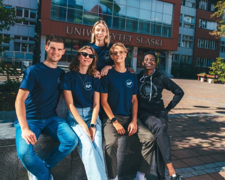 Pięciu młodych dorosłych siedzi i stoi razem, uśmiechając się przed budynkiem uniwersyteckim z napisem Uniwersytet Śląski. Mają na sobie luźne ubrania i niebieskie koszulki, ciesząc się słonecznym dniem na kampusie.