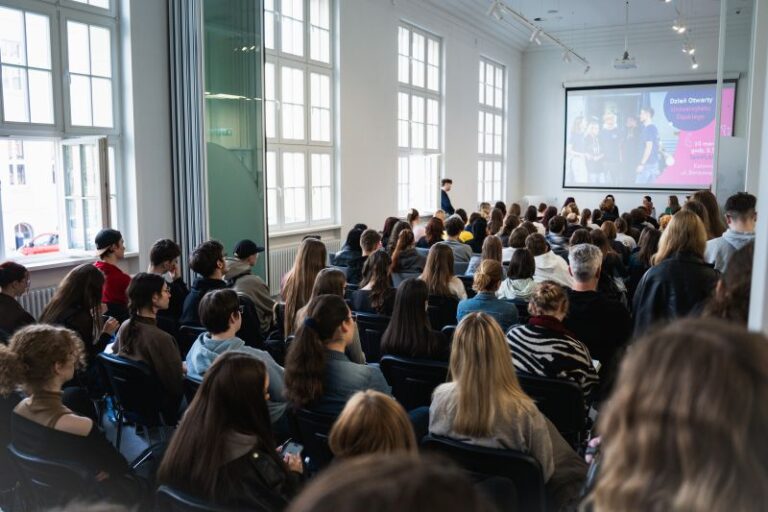 Duża grupa ludzi siedzi w rzędach naprzeciwko ekranu z przodu jasnego, nowoczesnego pokoju podczas prezentacji lub wykładu. Niektórzy uczestnicy uważnie obserwują, podczas gdy inni rozglądają się dookoła.