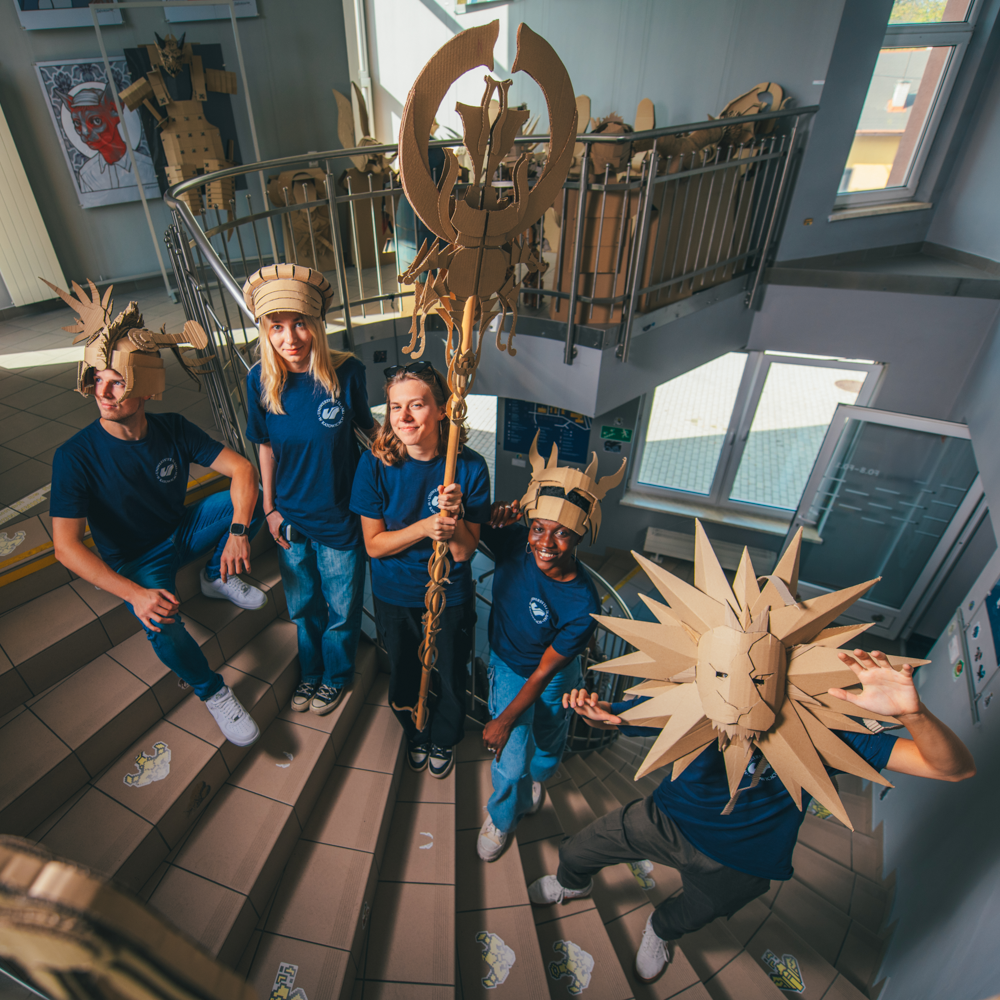 Cztery osoby w niebieskich koszulach i dżinsach stoją na wewnętrznych schodach, nosząc wyszukane kartonowe kapelusze i trzymając kartonowe rekwizyty, w tym dużą laskę i maskę w kształcie słońca. Tekturowe rzeźby są wyświetlane wokół obszaru.