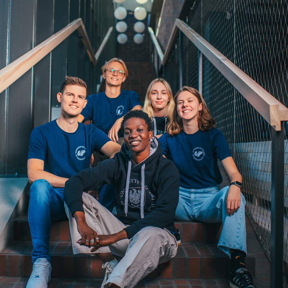 Pięciu młodych dorosłych siedzi na schodach, uśmiechając się do kamery. Czworo z nich ma na sobie pasujące do siebie niebieskie koszulki, a jeden z nich nosi czarną bluzę z kapturem. Sceneria jest nowoczesna, z metalową poręczą i wiszącymi światłami nad głową.
