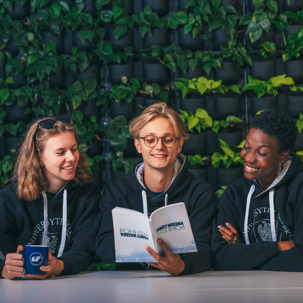 Trzech młodych dorosłych w bluzach siedzi przy stole, uśmiechając się i czytając razem książkę. Jeden z nich trzyma kubek. Bujna zielona ściana z roślinami służy jako tło, tworząc relaksującą i przyjazną atmosferę.