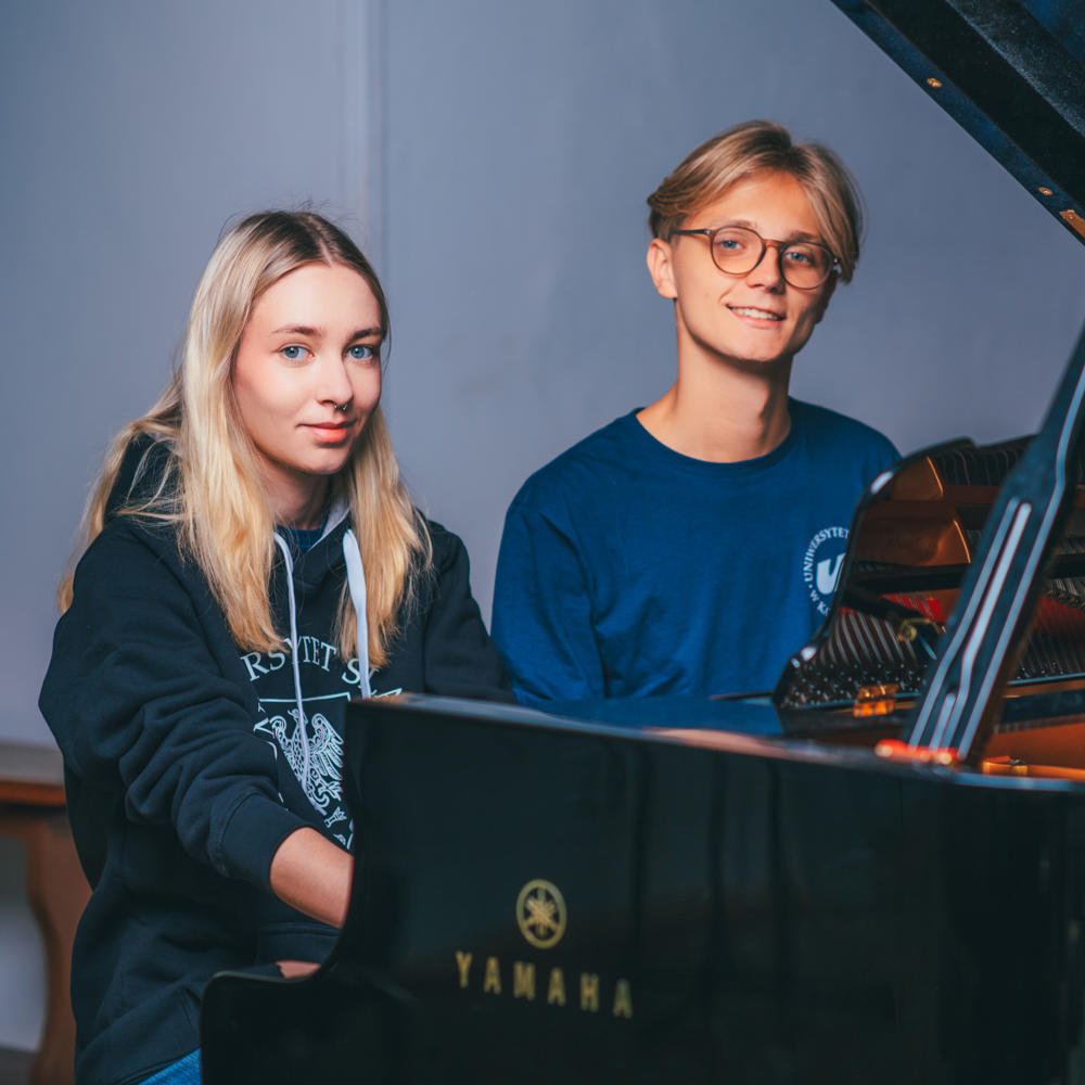 Dwoje młodych dorosłych, jeden z długimi blond włosami i jeden z krótkimi jasnymi włosami i okularami, siedzą razem przy czarnym fortepianie Yamaha, oboje patrzą w kamerę i uśmiechają się w delikatnie oświetlonym wnętrzu.