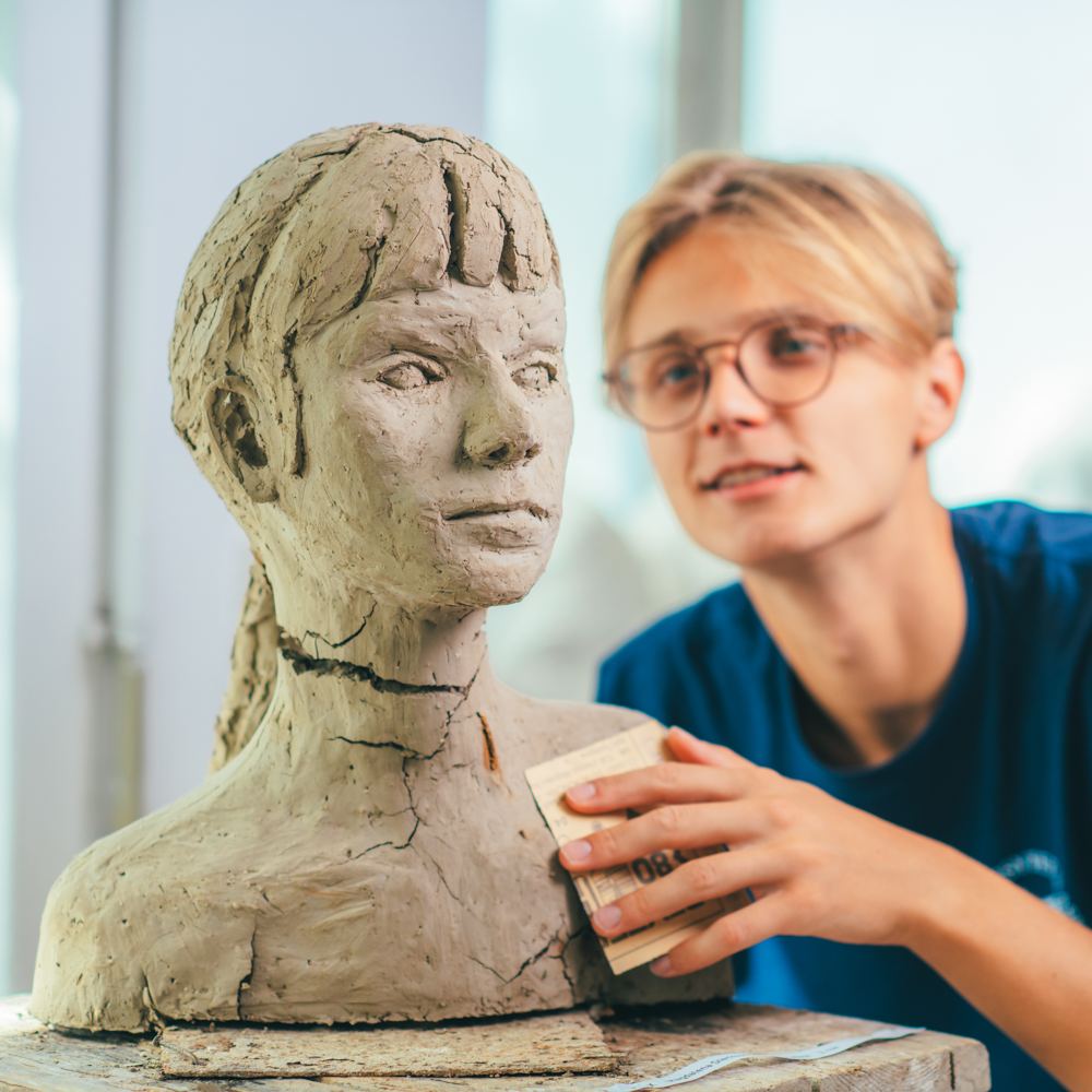 Młoda osoba w okularach kształtuje gliniane popiersie głowy kobiety, używając papieru ściernego do wygładzenia powierzchni rzeźby. Skupiają się na swojej pracy w dobrze oświetlonym studio.