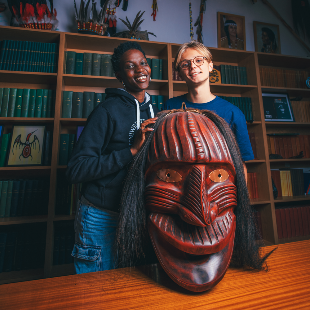 Dwie uśmiechnięte osoby stoją w bibliotece, a za nimi leżą kolorowe książki. Przed nimi znajduje się duża, czerwona, rzeźbiona drewniana maska o skomplikowanych liniach, ludzkich rysach i długich czarnych włosach.