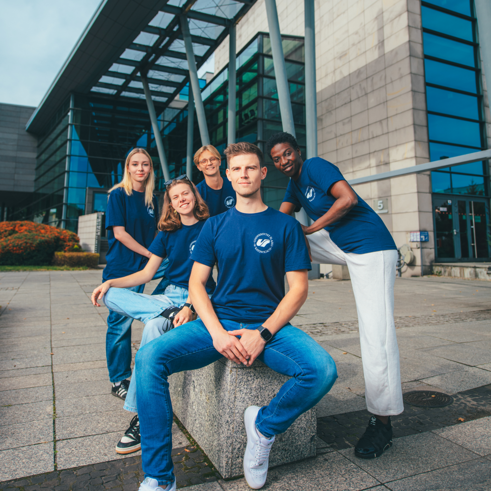Pięciu młodych dorosłych w dopasowanych niebieskich koszulach pozuje i uśmiecha się przed nowoczesnym budynkiem o szklano-betonowej architekturze. Czterech stoi z tyłu, a jeden siedzi na kamiennej ławce z przodu. Jest słoneczny dzień.
