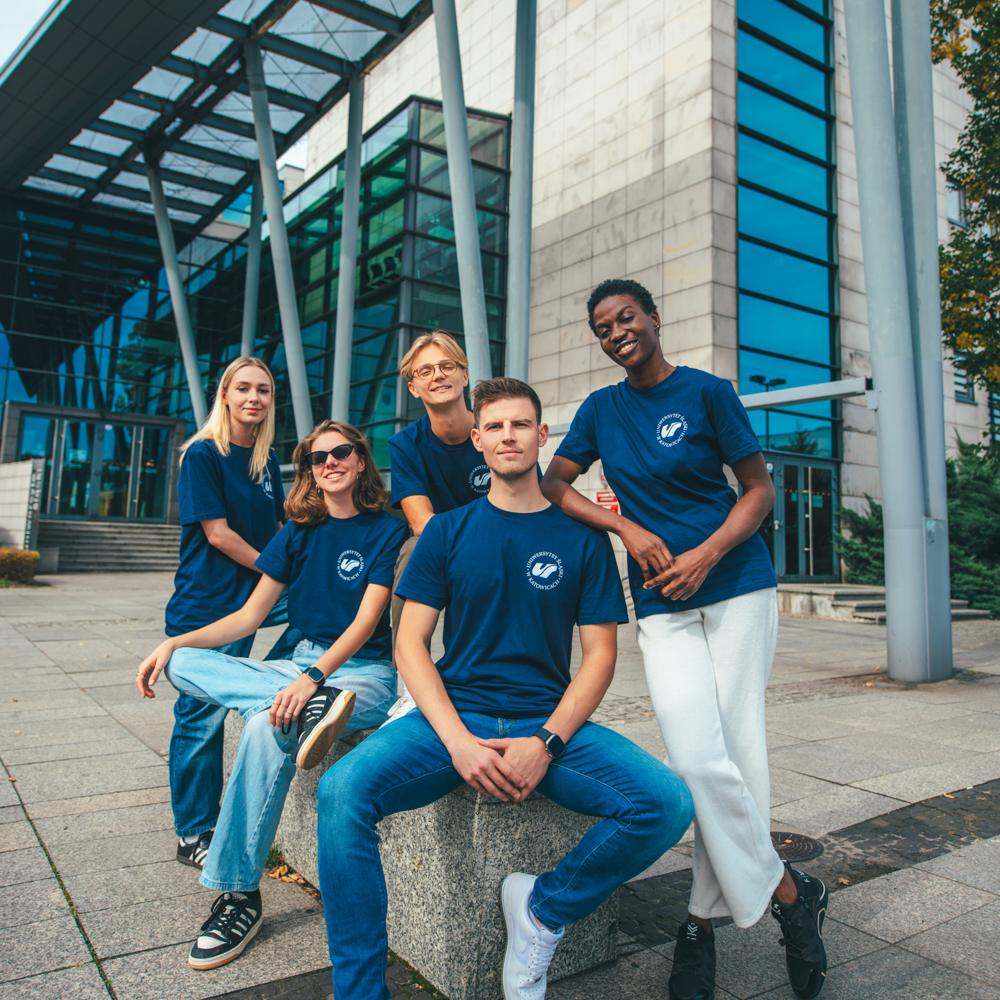 Pięć młodych osób ubranych w pasujące do siebie granatowe koszulki z białym okrągłym logo pozuje razem przed nowoczesnym budynkiem ze szkła i kamienia, uśmiechając się do kamery.