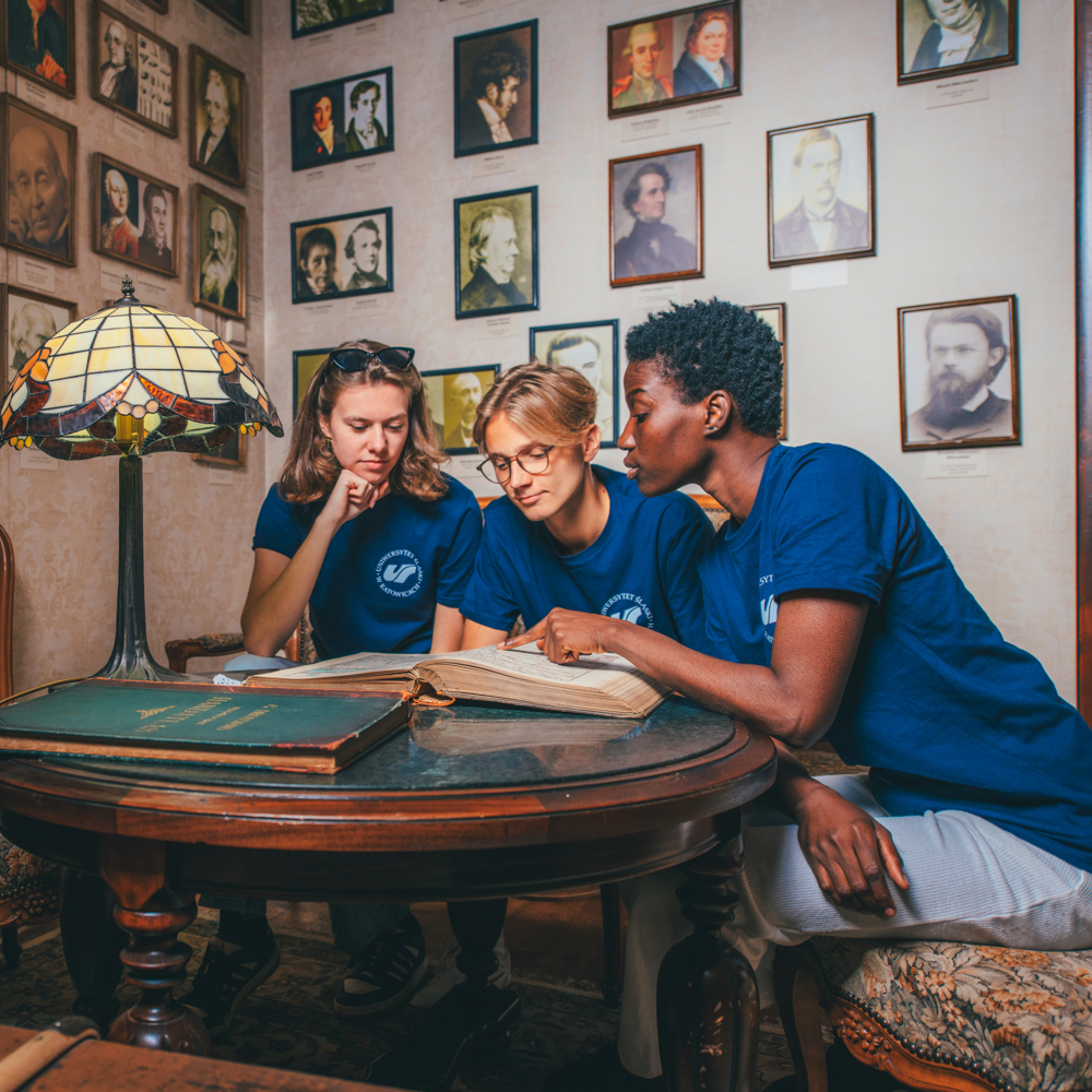 Trzy osoby w dopasowanych niebieskich koszulach siedzą wokół okrągłego drewnianego stołu w pokoju z oprawionymi portretami na ścianie, czytając razem otwartą książkę pod witrażową lampą.