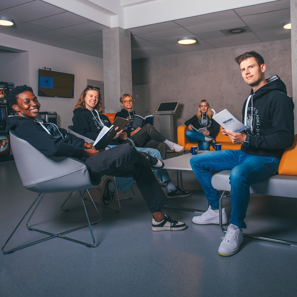 Pięć osób siedzi razem w nowoczesnym salonie, swobodnie ubranych i trzymających broszury. Są uśmiechnięci i wydają się prowadzić swobodną dyskusję grupową. Otoczenie jest jasne z wygodnymi miejscami do siedzenia.