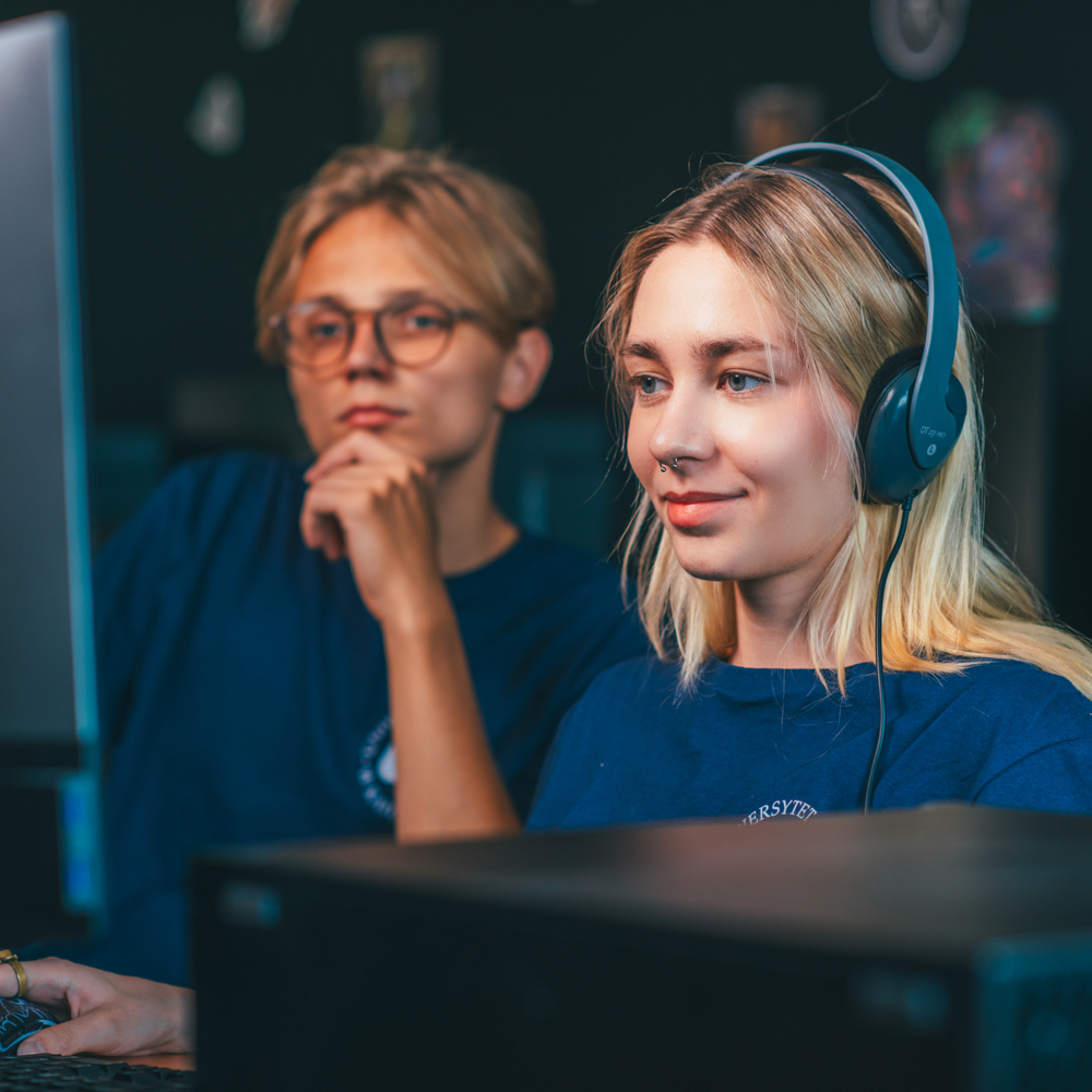 Dwoje młodych ludzi w niebieskich koszulach siedzi przy monitorach komputerów. Kobieta z przodu nosi słuchawki i uśmiecha się, podczas gdy mężczyzna za nią wygląda na zamyślonego z ręką na brodzie. Oboje wydają się skupieni na ekranach.