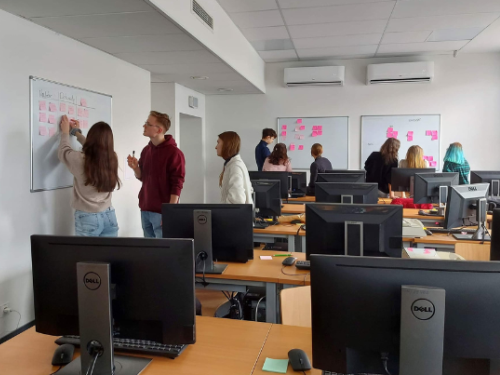 Grupa studentów w pracowni komputerowej pracuje razem, pisząc na tablicach pokrytych karteczkami samoprzylepnymi, podczas gdy inni dyskutują lub obserwują. Monitory komputerowe są widoczne na biurkach na pierwszym planie.