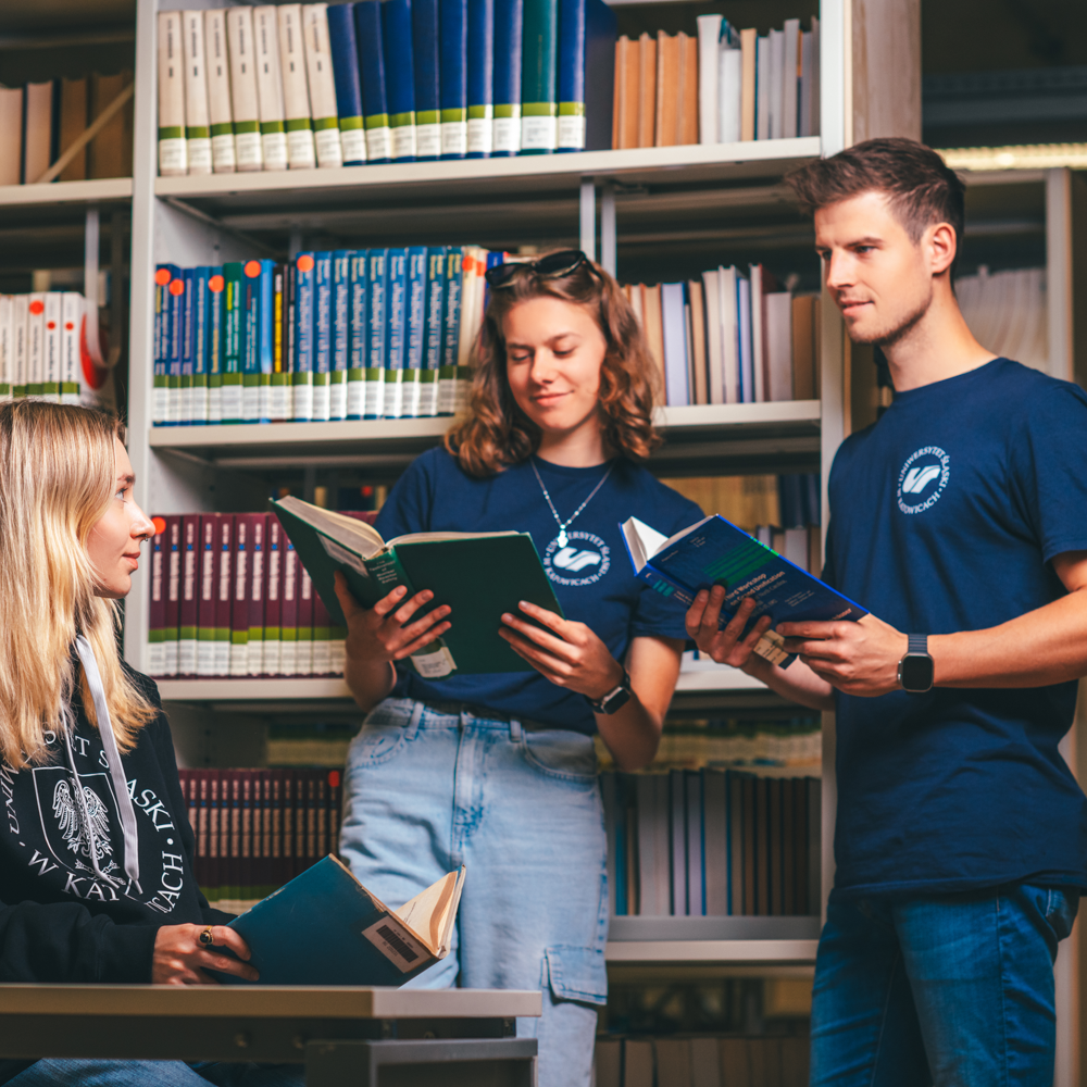 Trzech młodych dorosłych w swobodnych ubraniach zbiera się w bibliotece, trzymając książki i czytając razem. W tle widać półki wypełnione książkami, a jedna osoba siedzi przy biurku, podczas gdy pozostałe stoją.