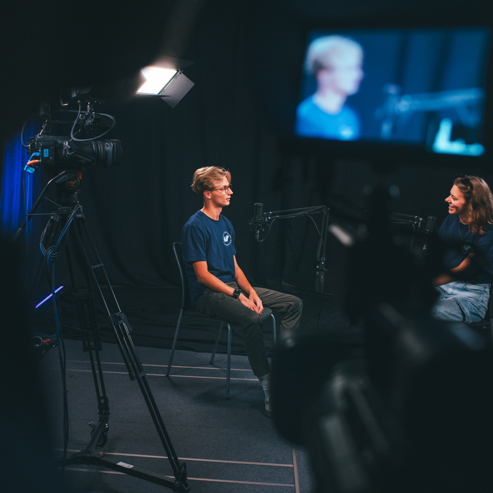 Dwie osoby siedzą naprzeciwko siebie w ciemnym studiu, otoczone kamerami wideo, mikrofonami i jasnymi światłami, nagrywając coś, co wydaje się być wywiadem lub podcastem.