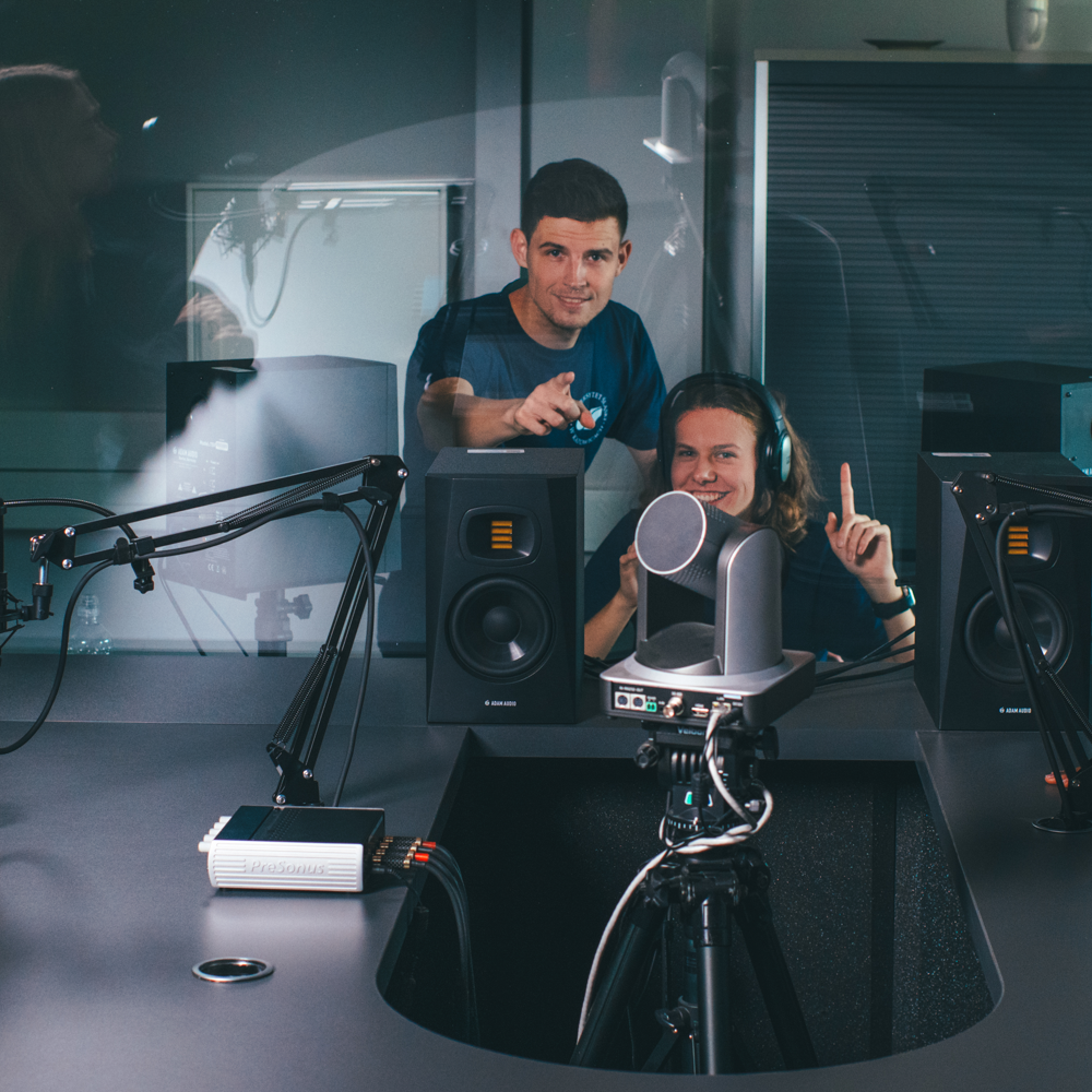 Dwie osoby w studiu nagraniowym siedzą za szklaną szybą, noszą słuchawki i uśmiechają się. Jedna z nich wskazuje na kamerę, a druga unosi palec. Na pierwszym planie widoczne są mikrofony, głośniki i sprzęt audio.