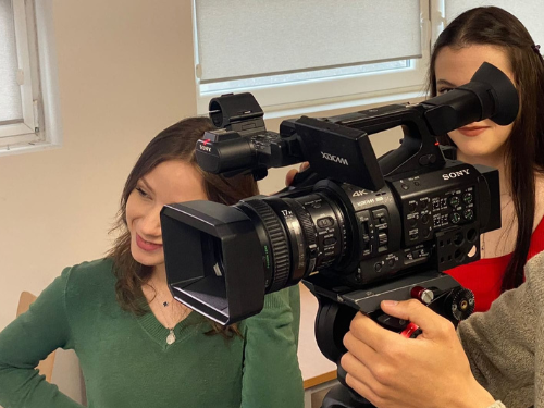 Dwie kobiety, jedna w zielonej, a druga w czerwonej bluzce, stoją w pomieszczeniu obok dużej profesjonalnej kamery wideo Sony 4K na statywie. Kamera jest obsługiwana przez kogoś poza kadrem.