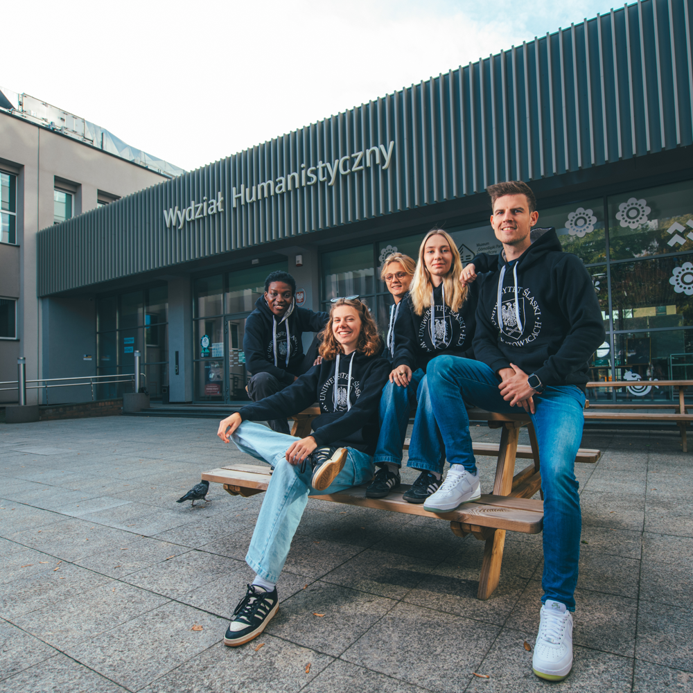 Pięcioro młodych ludzi w dopasowanych czarnych bluzach siedzi i pozuje na drewnianej ławce przed budynkiem Wydziału Humanistycznego. Są uśmiechnięci, a pogoda jest bezchmurna.
