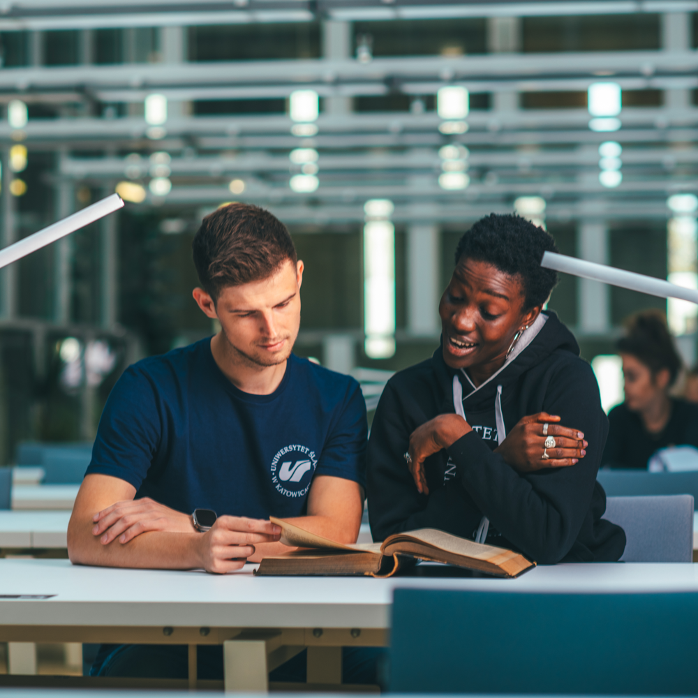 Dwóch studentów siedzi przy stole w jasnej bibliotece, studiując razem z otwartą książką. Jeden z nich czyta, podczas gdy drugi gestykuluje i entuzjastycznie coś wyjaśnia.
