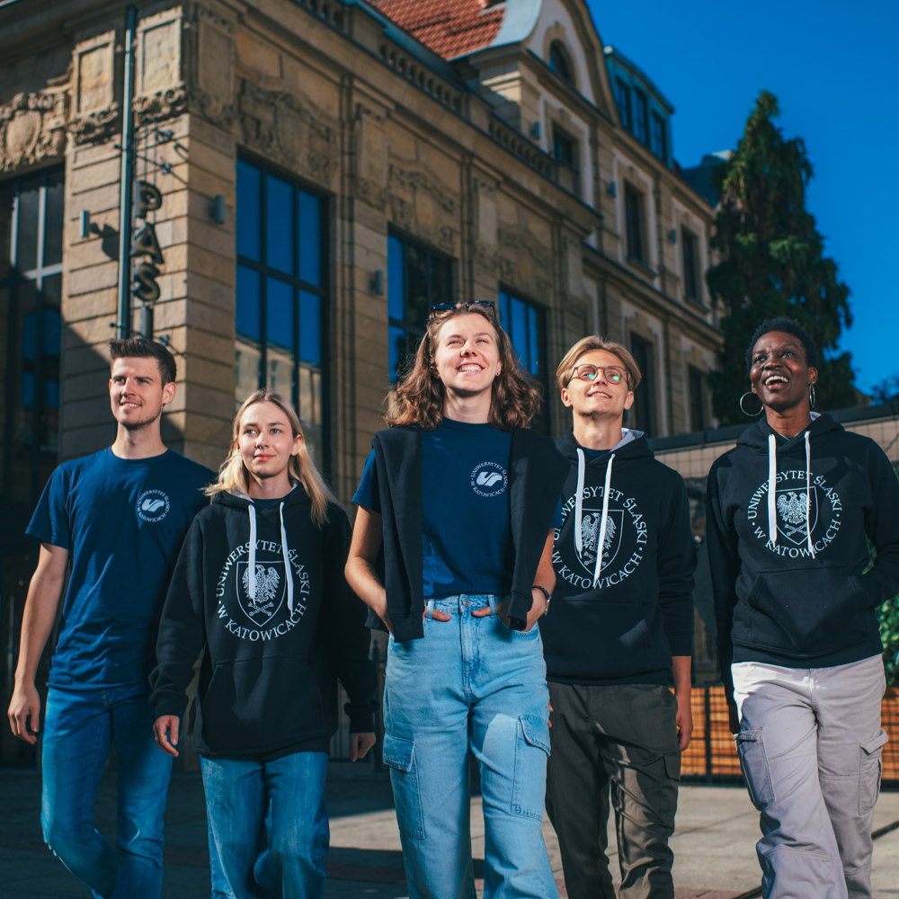 Pięciu młodych dorosłych stoi na zewnątrz, uśmiechając się i spacerując razem w swobodnych strojach i markowych ubraniach uniwersyteckich, przed zabytkowym budynkiem pod jasnym, błękitnym niebem.