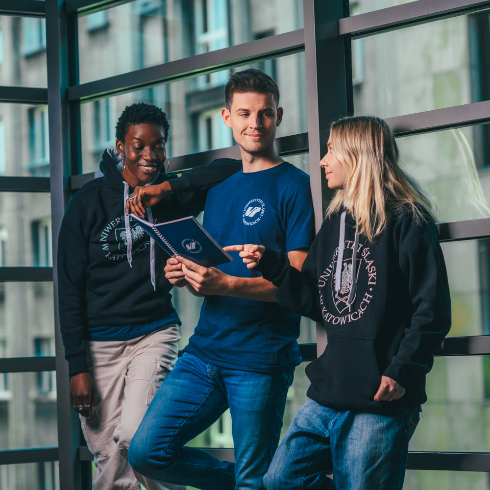 Trzech młodych dorosłych stoi w pomieszczeniu przy dużym oknie, uśmiechając się i patrząc razem na dokument. Mają na sobie zwykłe dżinsy i bluzy z nadrukiem Uniwersytetu Śląskiego w Katowicach.