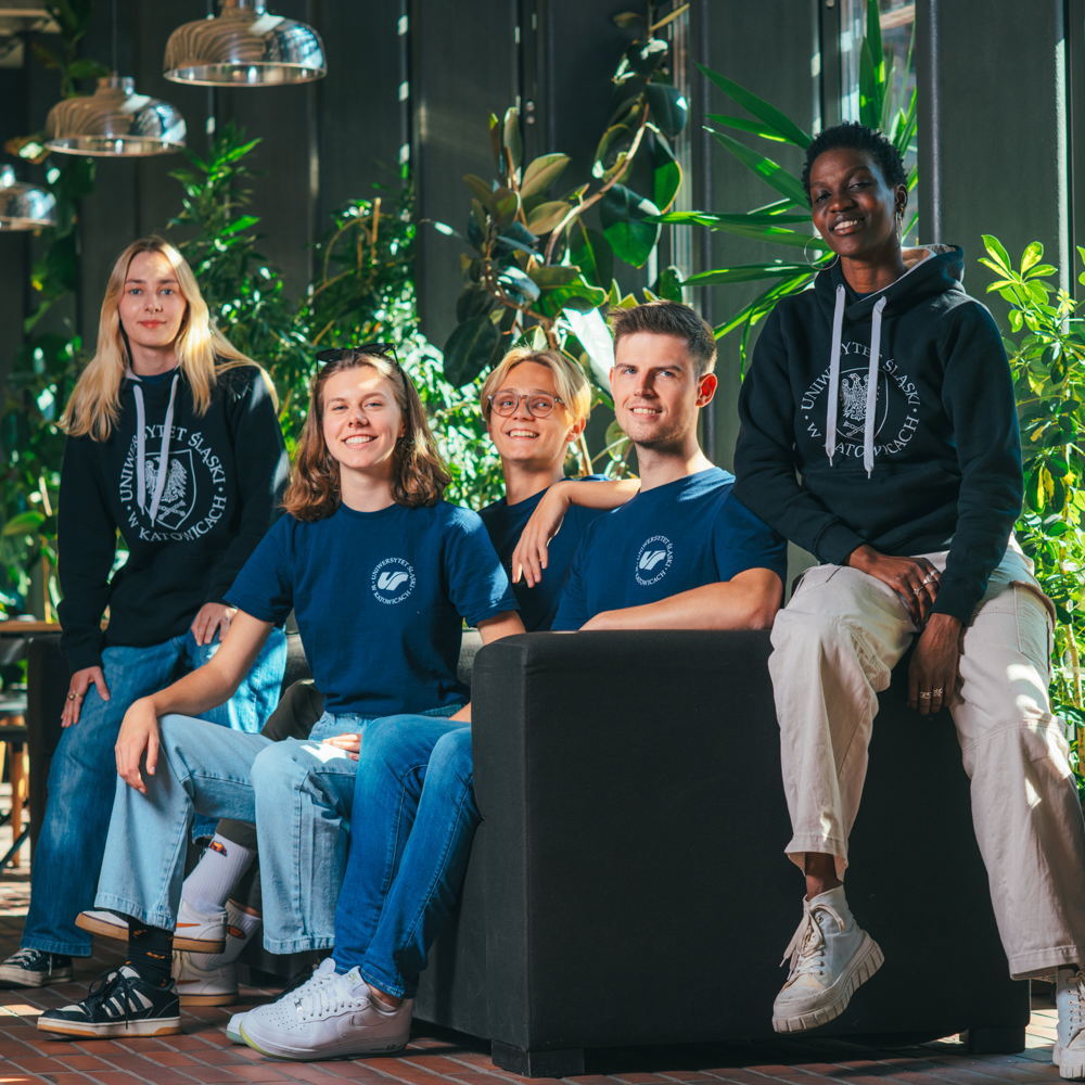 Pięć osób siedzi razem w jasnym pomieszczeniu z wieloma zielonymi roślinami. Cztery siedzą na sofie, a jedna siedzi na podłokietniku. Uśmiechają się do kamery, ubrani w pasujące do siebie niebieskie koszulki i bluzy z kapturem.