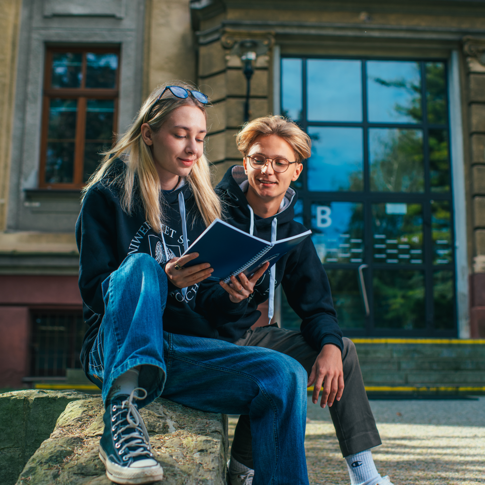 Dwóch studentów siedzi przed budynkiem, jeden czyta z notatnika, podczas gdy drugi słucha. Obaj noszą swobodne ubrania i bluzy z kapturem, wyglądając na zaangażowanych i zrelaksowanych w otoczeniu kampusu.