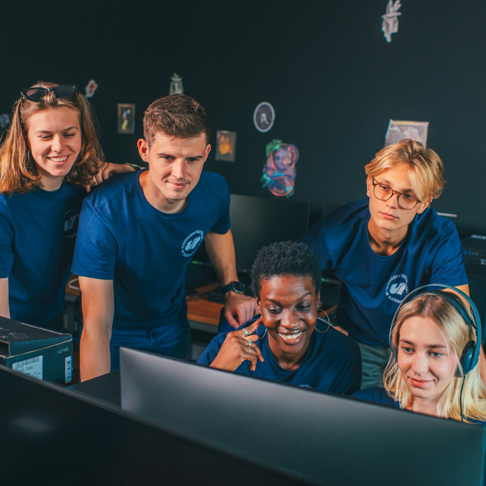 Pięć młodych osób w pasujących do siebie niebieskich koszulach zebrało się wokół monitora komputera, uśmiechając się i wyglądając na zaangażowanych, co sugeruje pracę zespołową lub współpracę w nowoczesnym biurze lub klasie.