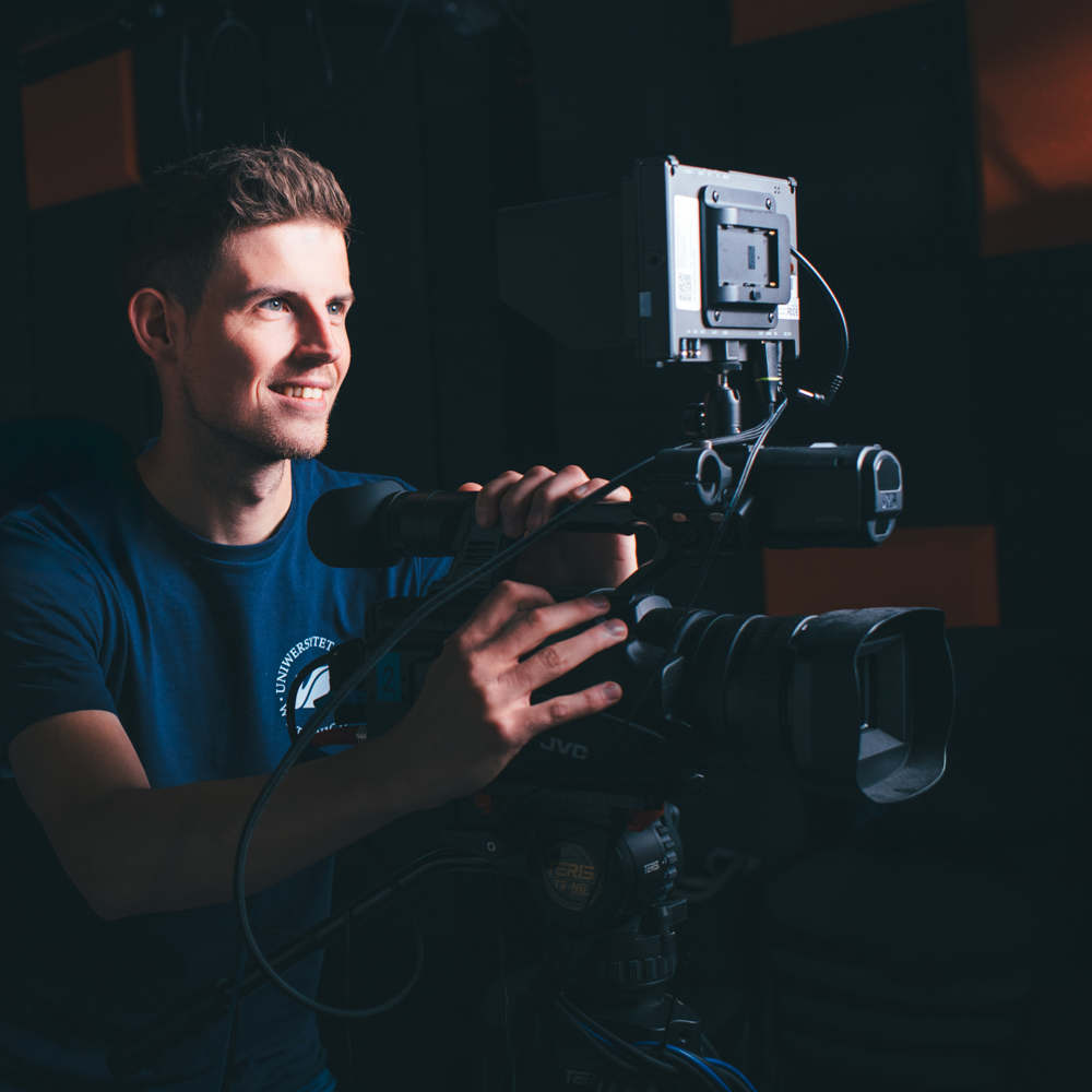 Uśmiechnięty mężczyzna w ciemnej koszulce obsługuje profesjonalną kamerę wideo w słabo oświetlonym studiu.