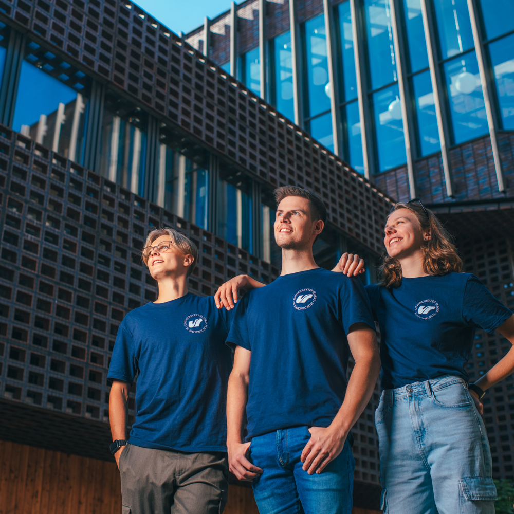Trzech młodych dorosłych ubranych w pasujące do siebie niebieskie koszulki z białym logo stoi obok siebie na zewnątrz, uśmiechając się i patrząc w górę, z nowoczesnymi szklanymi i ceglanymi budynkami w tle.