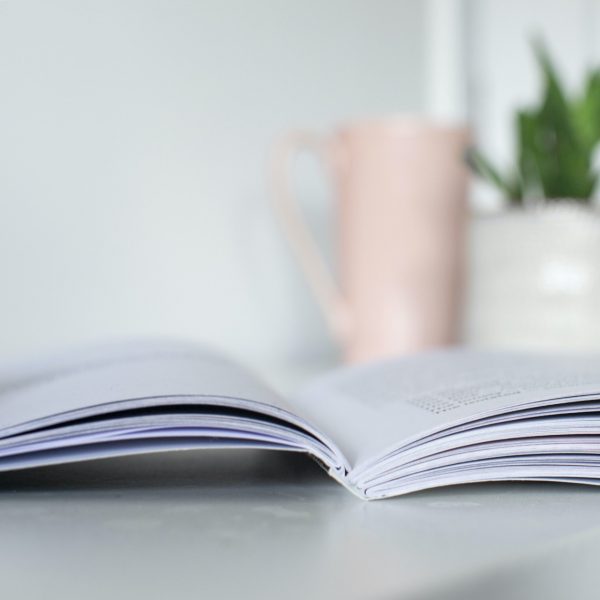 Otwarta książka leży na jasnej powierzchni, z rozmytym różowym kubkiem i rośliną doniczkową w tle, tworząc spokojną i przytulną atmosferę.