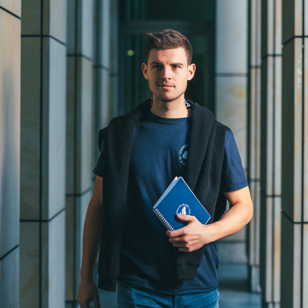 Młody mężczyzna z krótkimi brązowymi włosami idzie korytarzem wyłożonym kolumnami, ubrany w granatową koszulkę i trzymający niebieski spiralny notatnik. Ma czarną bluzę narzuconą na ramiona i pewnie patrzy w kamerę.