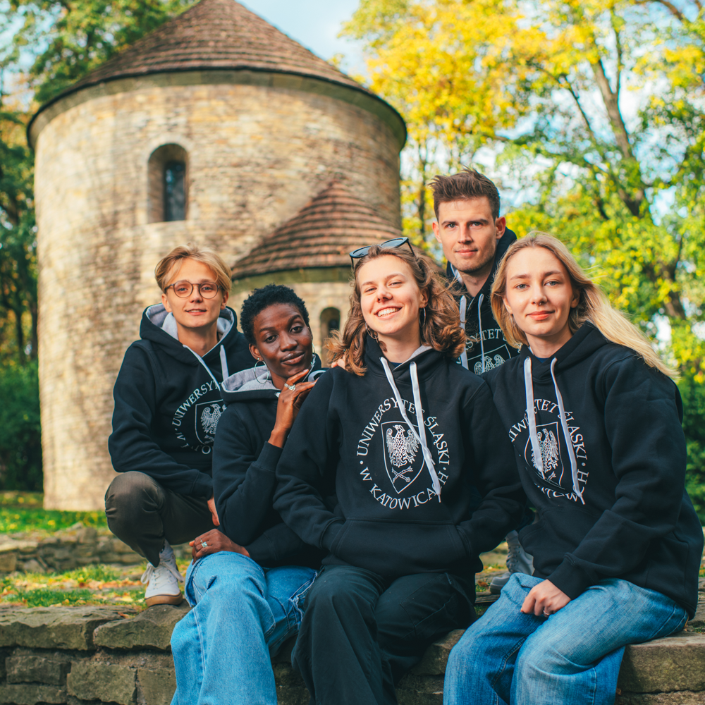 Pięć młodych osób ubranych w czarne bluzy z kapturem Uniwersytetu Śląskiego w Katowicach siedzi i pozuje razem, uśmiechając się, przed okrągłym kamiennym budynkiem otoczonym drzewami i zielenią.