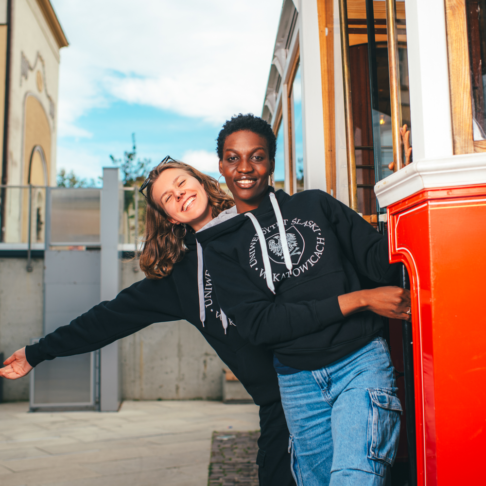 Dwie osoby ubrane w czarne bluzy z kapturem uśmiechają się i wychylają z drzwi jaskrawoczerwonego tramwaju, ciesząc się słonecznym dniem na świeżym powietrzu z nowoczesnymi budynkami w tle.