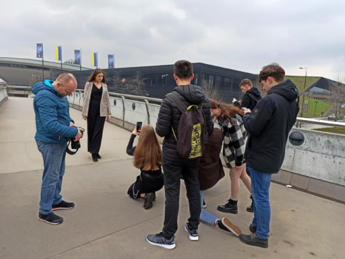 Kobieta pozuje na moście, podczas gdy kilka osób robi jej zdjęcia telefonami. Inni stoją w pobliżu, niektórzy z plecakami i sprzętem, pod zachmurzonym niebem z budynkami w tle.