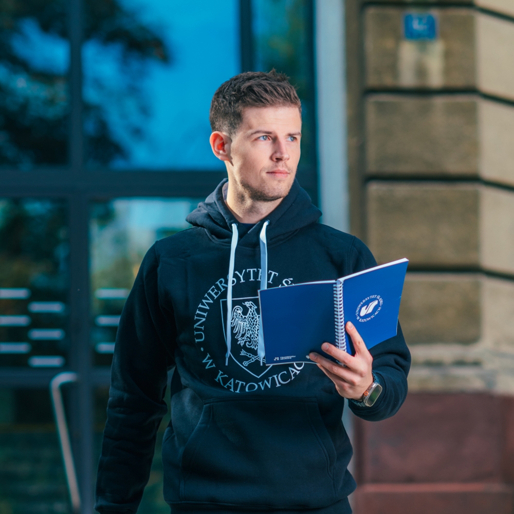 Młody mężczyzna ubrany w czarną bluzę z kapturem Uniwersytetu Śląskiego w Katowicach stoi na zewnątrz, trzymając otwarty niebieski notatnik, z budynkiem i szklanymi drzwiami w tle.