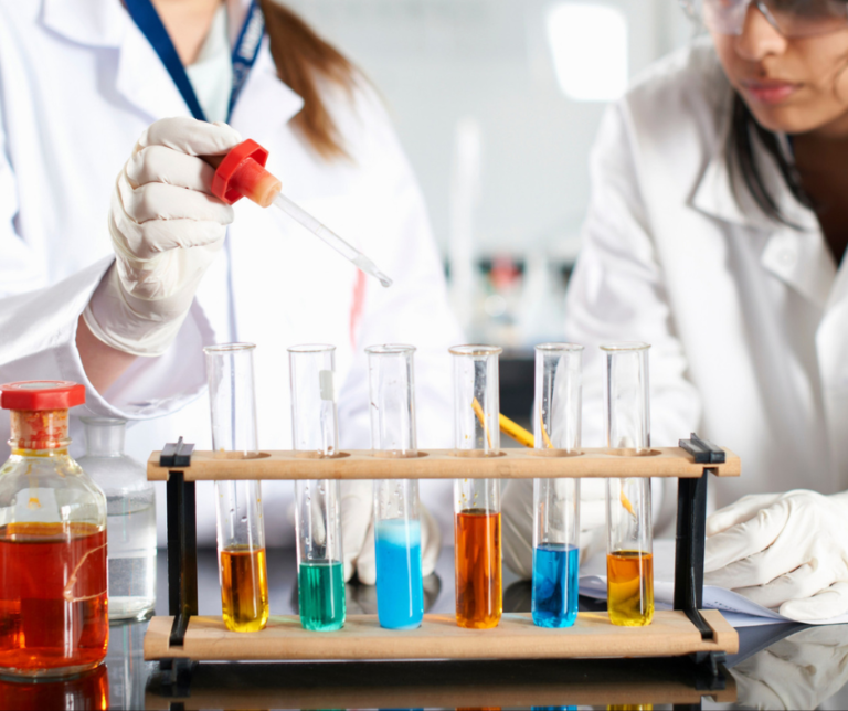Dwie osoby w fartuchach laboratoryjnych i rękawiczkach pracują z probówkami zawierającymi kolorowe płyny na stole laboratoryjnym. Jedna z nich używa zakraplacza, widoczne są też różne butelki i sprzęt naukowy.