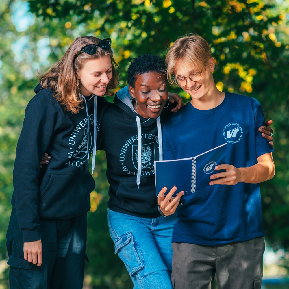 Trzech uśmiechniętych studentów stoi na zewnątrz, patrząc razem na notatnik. Dwóch z nich ma na sobie czarne bluzy uniwersyteckie, a jeden nosi niebieską koszulkę uniwersytecką. W tle zielone drzewa i światło słoneczne.