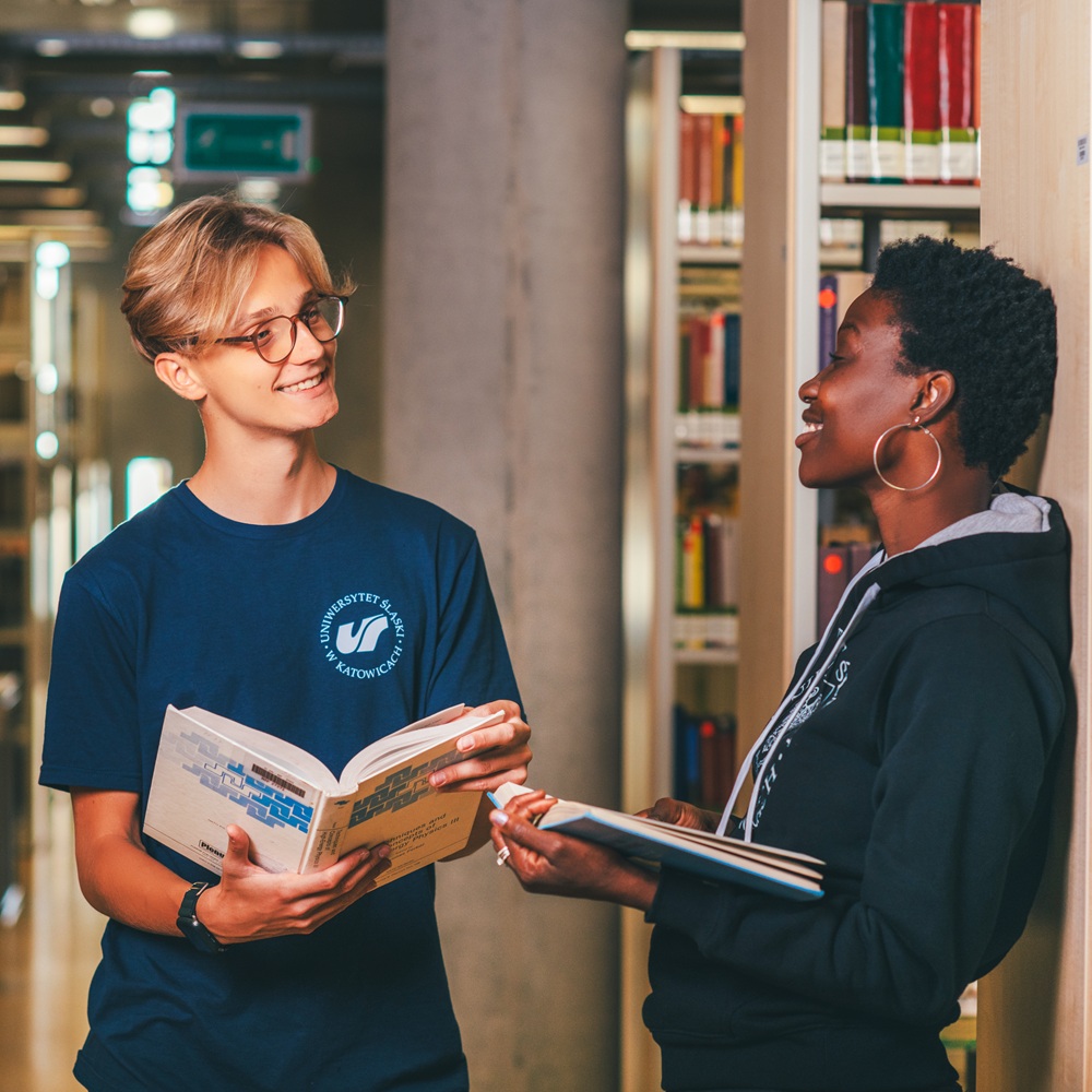 Dwóch studentów stoi w bibliotece, uśmiechając się i rozmawiając, trzymając otwarte książki. Jeden z nich nosi okulary i niebieską koszulkę, a drugi kolczyki w uszach i czarną bluzę z kapturem. Za nimi widoczne są półki z książkami.