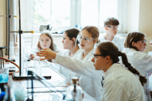 Grupa studentów ubranych w fartuchy laboratoryjne i okulary ochronne wspólnie przeprowadza eksperymenty naukowe przy stole laboratoryjnym, na którym leżą naczynia szklane i sprzęt.