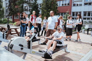 Dwóch chłopców korzysta z wioślarzy na świeżym powietrzu, podczas gdy grupa osób, w tym dorośli i nastolatkowie, obserwuje ich i dopinguje. Akcja rozgrywa się na utwardzonym terenie w pobliżu stołów piknikowych i wielopiętrowego budynku.