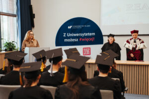 Uroczystość wręczenia dyplomów na Uniwersytecie Śląskim, podczas której studenci w czapkach i togach stoją twarzą do sceny, na której trzech wykładowców – z których dwóch ma na sobie togi akademickie – stoi obok tablicy z napisem „Z Uniwersytetem możesz #więcej!”.