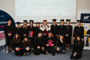 Grupa studentów w czarnych togach i czapkach absolwentów pozuje w pomieszczeniu wraz z osobą dorosłą ubraną w uroczyste szaty i czapkę. Stoją przed ekranem, na którym widnieje napis „REAKCJI” oraz sztandary uniwersyteckie.