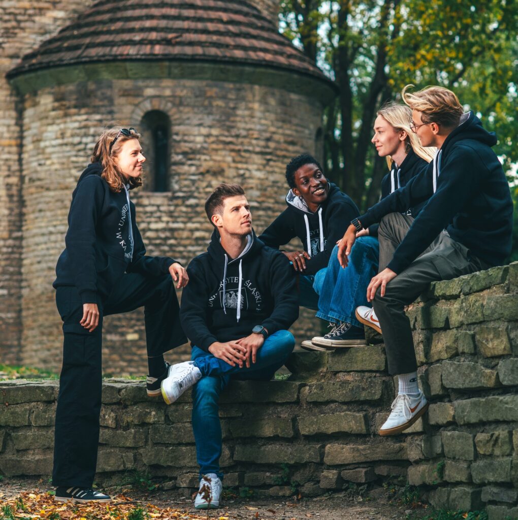Pięć osób ubranych w identyczne czarne bluzy z kapturem siedzi i stoi na kamiennych schodach na świeżym powietrzu, uśmiechając się i rozmawiając w pobliżu starego ceglanego budynku otoczonego drzewami.