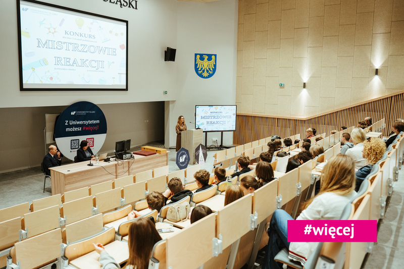 Kobieta przemawia z podium w nowoczesnej sali wykładowej, w której publiczność siedzi w rzędach. Na slajdzie prezentacji widnieje napis „Konkurs Mistrzowie Reakcji”. Widoczny jest baner oraz dwóch panelistów. W prawym dolnym rogu znajduje się hashtag #więcej.